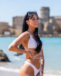 Donna sorridente in bikini bianco con capelli neri in spiaggia, con castello antico sfocato sullo sfondo.