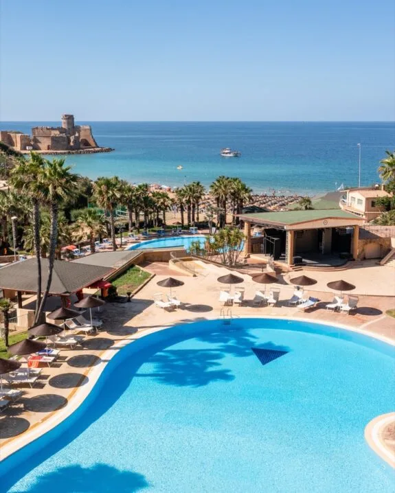 Piscina di hotel con ombrelloni, palme, spiaggia e mare con castello sullo sfondo in una giornata soleggiata.