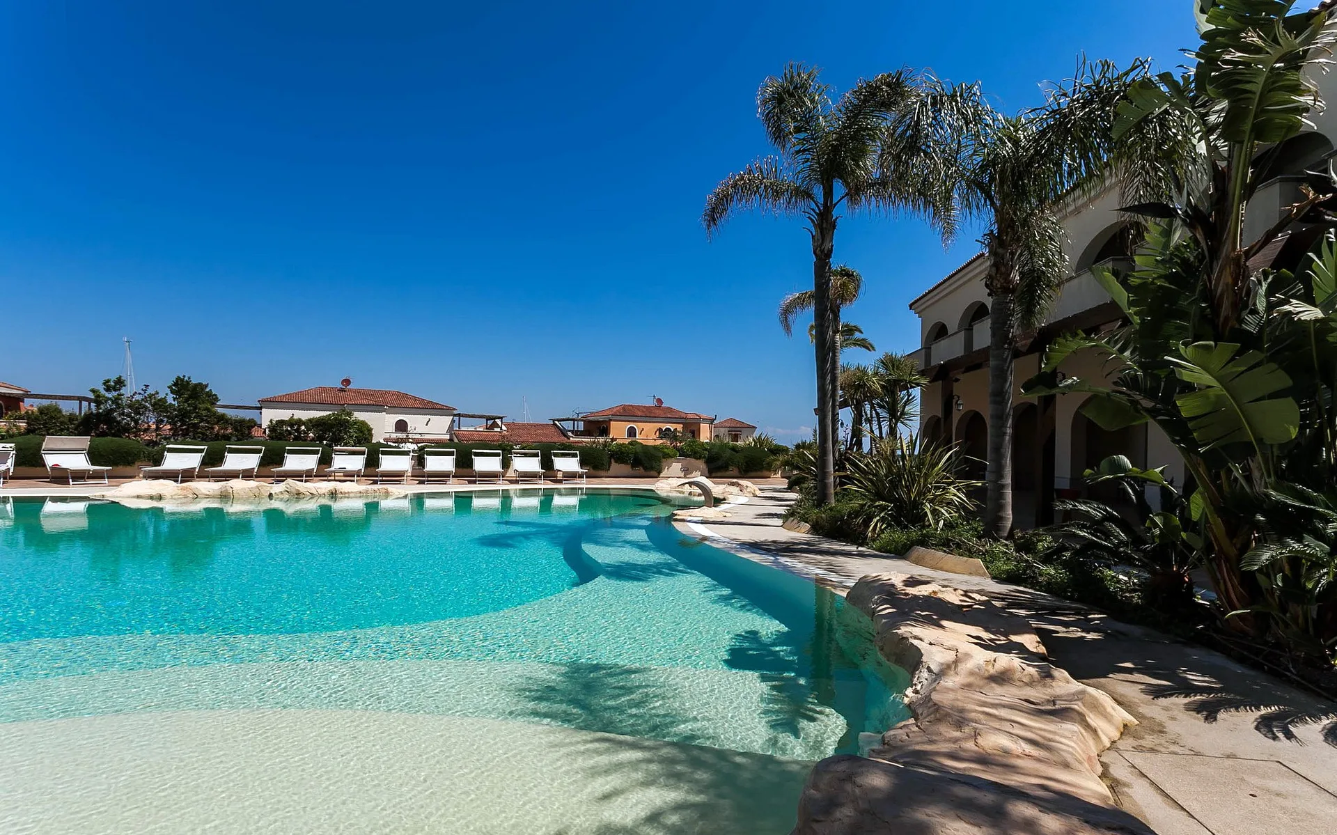 Piscina esterna con ingresso a spiaggia, lettini bianchi e palme accanto agli edifici