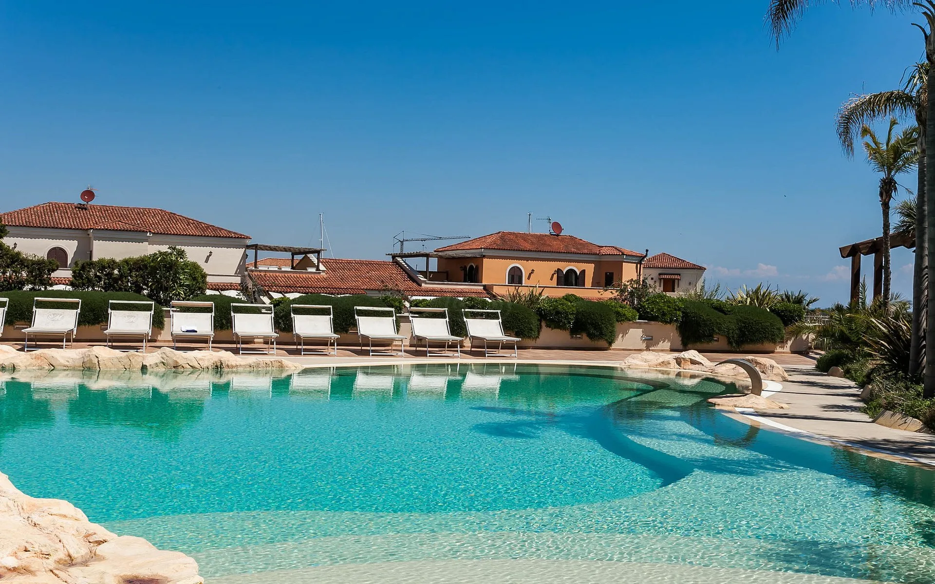 Piscina esterna con lettini bianchi allineati e edifici del resort con tetti in tegole