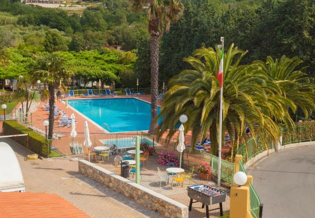 Vista sulla piscina con palme, lettini blu e area relax con tavolini e calcio balilla