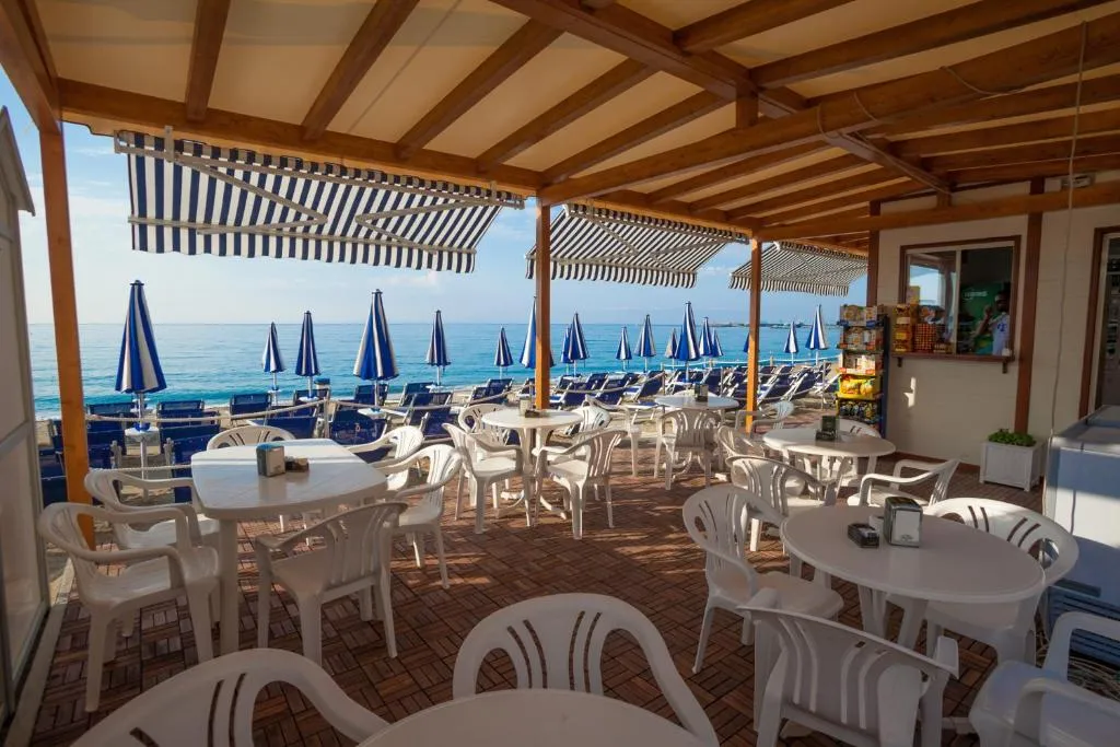 Terrazza bar sulla spiaggia con tavoli bianchi, pergolato in legno e vista sul mare con ombrelloni