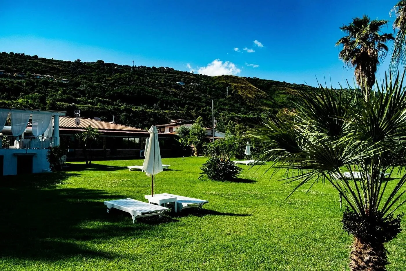 Giardino con prato verde, lettini bianchi e ombrelloni, colline sullo sfondo