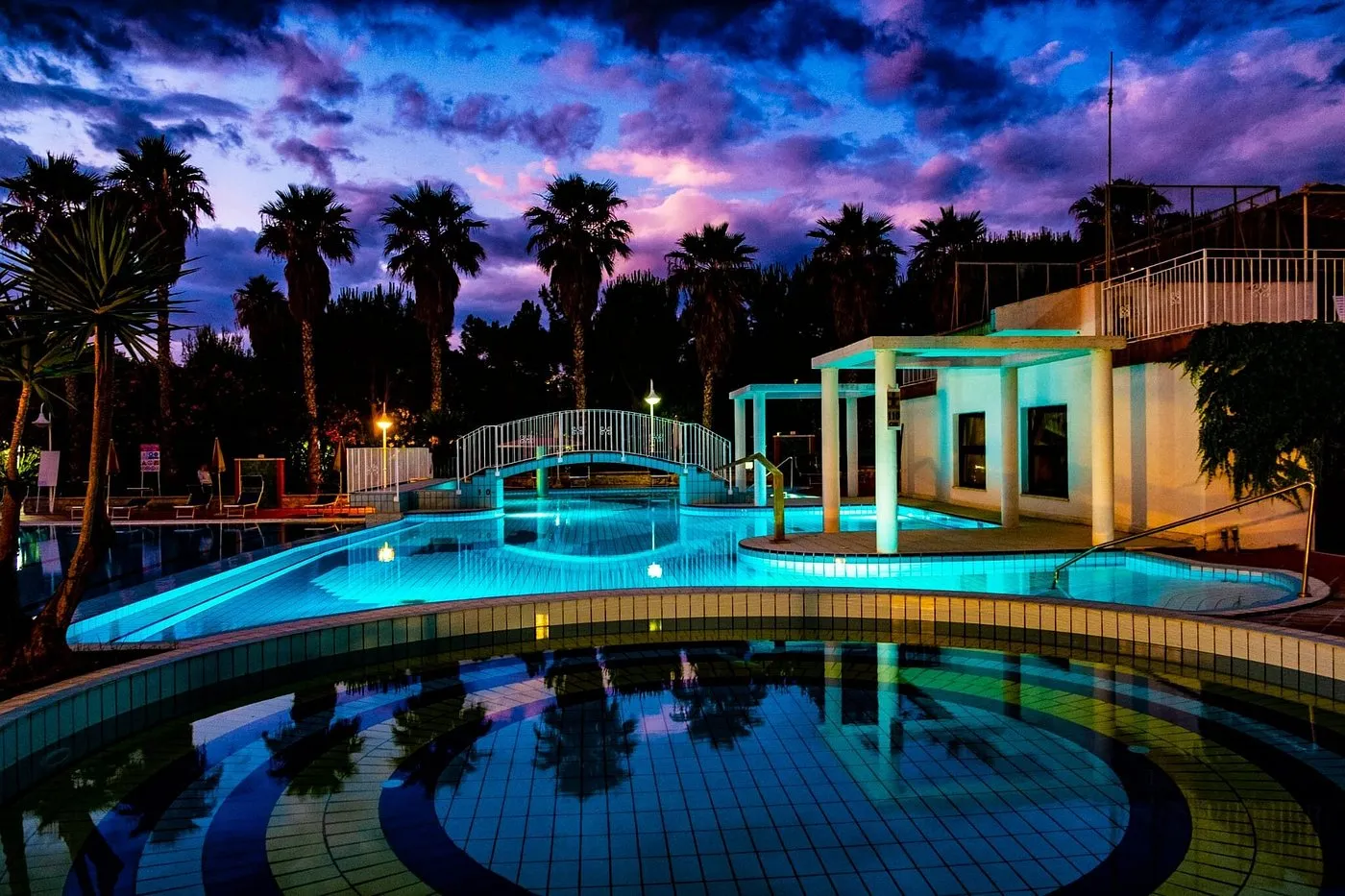 Piscina illuminata di notte con ponticello e cielo viola al crepuscolo