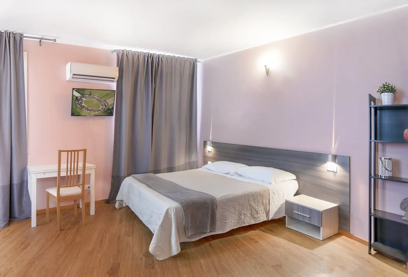 Camera con letto matrimoniale, scrivania bianca, condizionatore e mensole, dai toni rosa e grigi