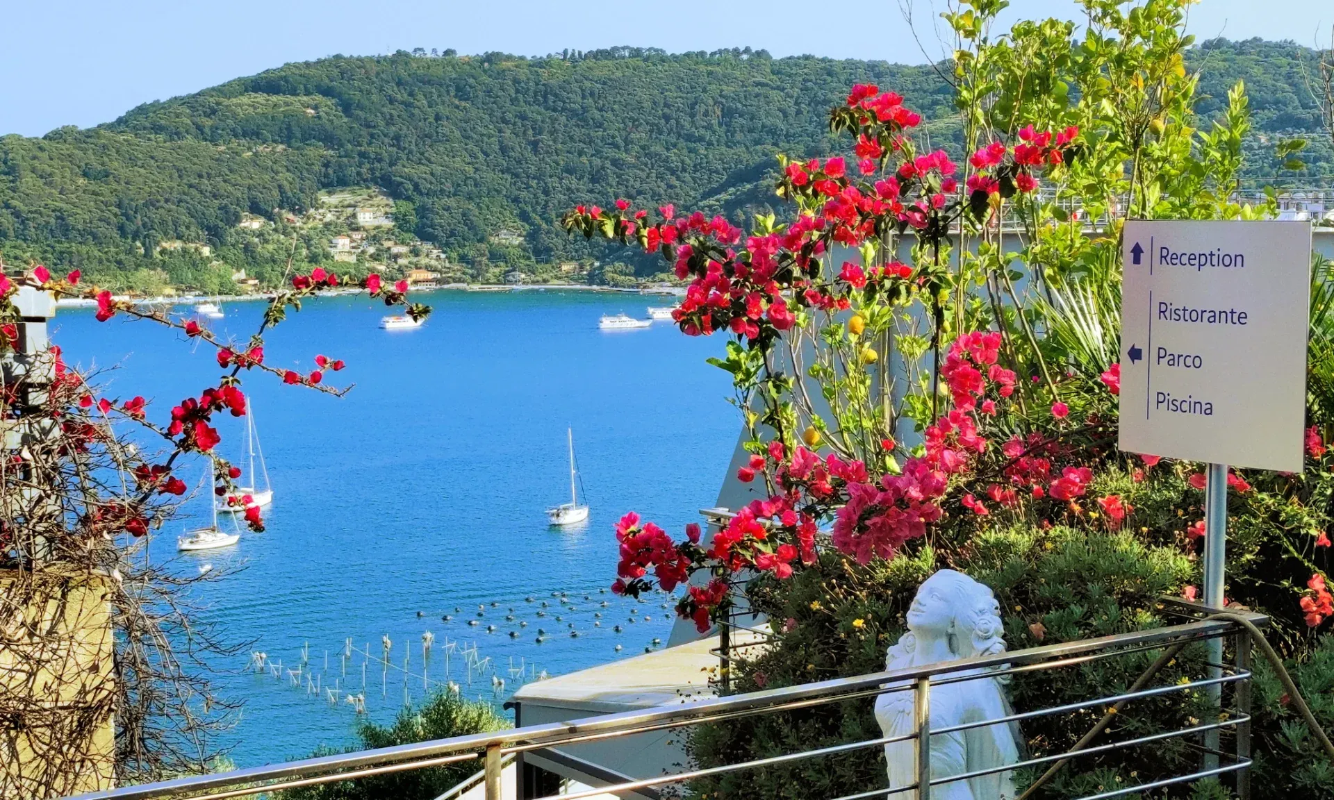 Vista sul golfo con barche a vela, bougainville fiorite e cartello indicazioni hotel