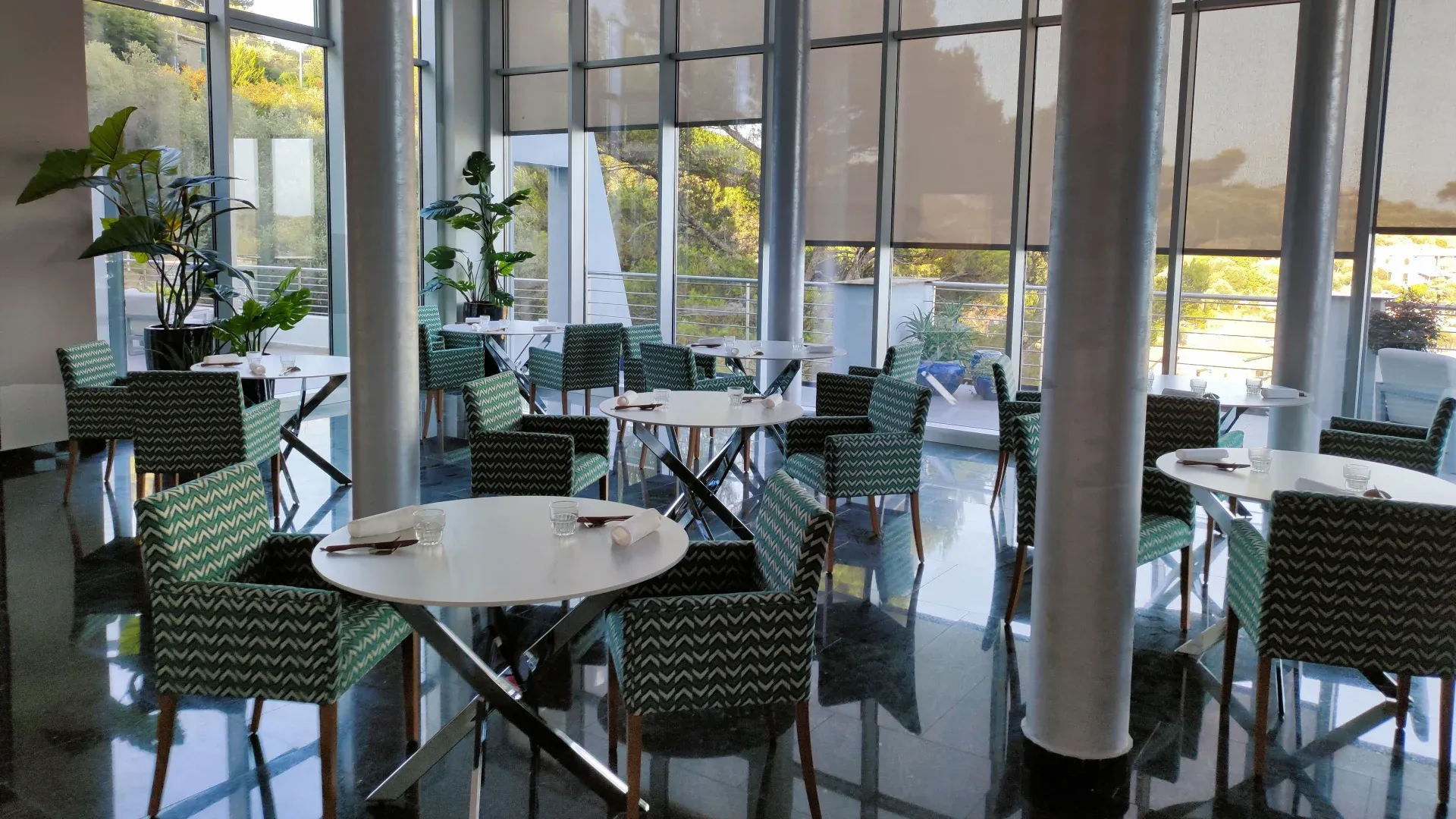 Sala ristorante luminosa con grandi vetrate, tavoli rotondi e sedie verdi a fantasia