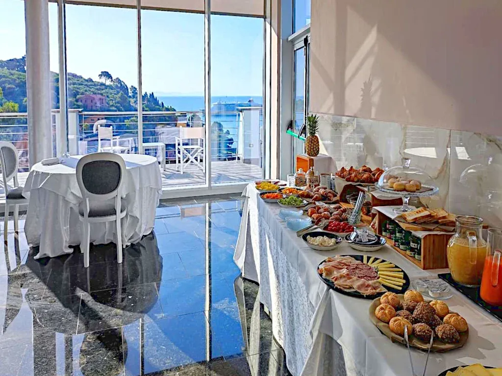 Buffet della colazione con dolci, frutta e salumi davanti a vetrate con vista mare