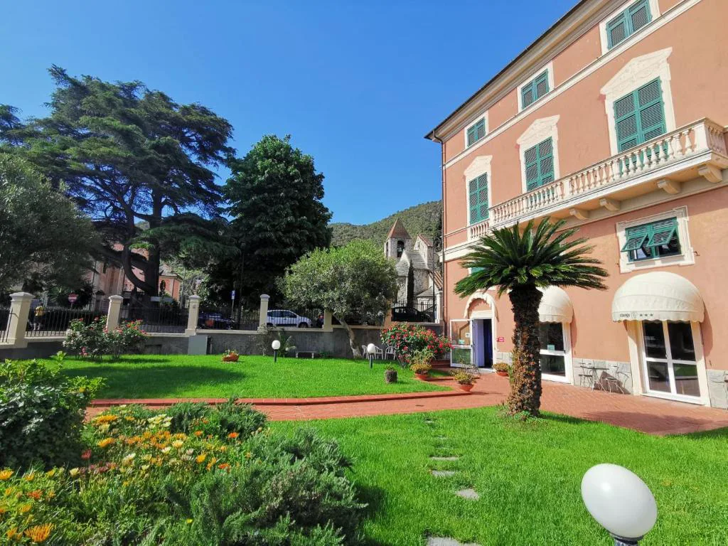 Giardino dell'hotel con prato verde, palma, aiuole fiorite e vista sulla villa storica e chiesetta