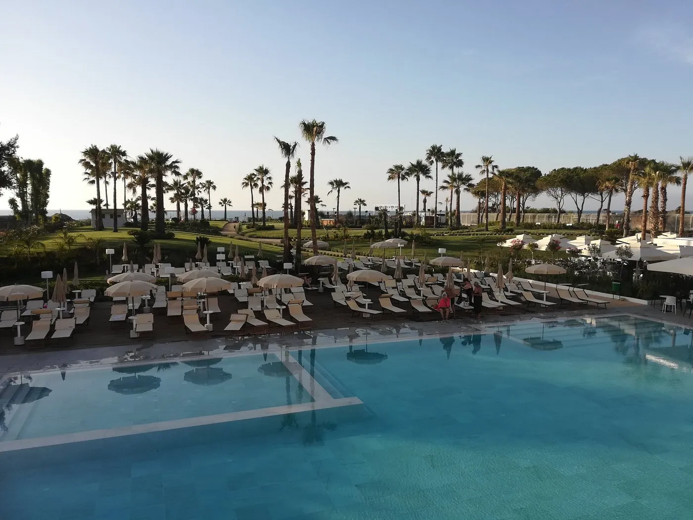 Piscina con lettini e ombrelloni al tramonto, palme e mare sullo sfondo