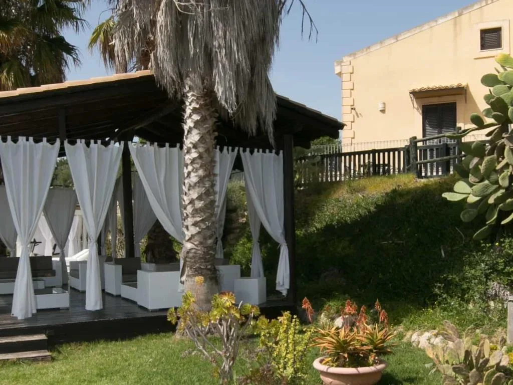 Gazebo in legno con tende bianche e lettini, circondato da palme e giardino con piante mediterranee