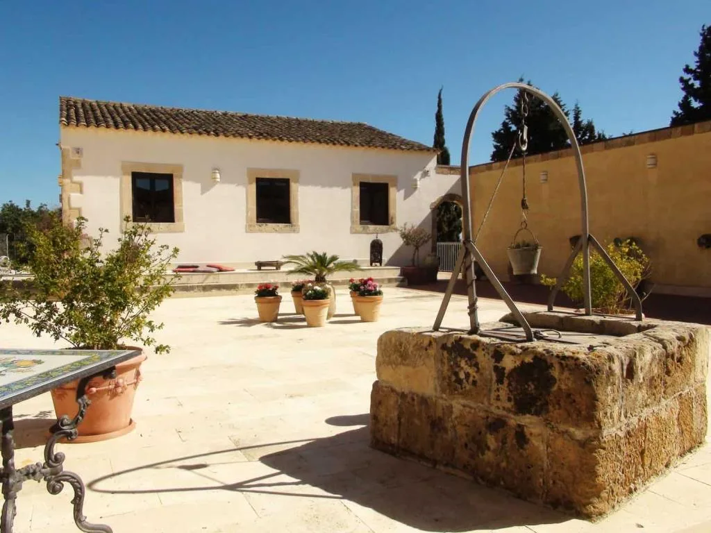 Cortile in pietra con antico pozzo, fioriere di gerani e edificio rustico bianco sotto cielo azzurro