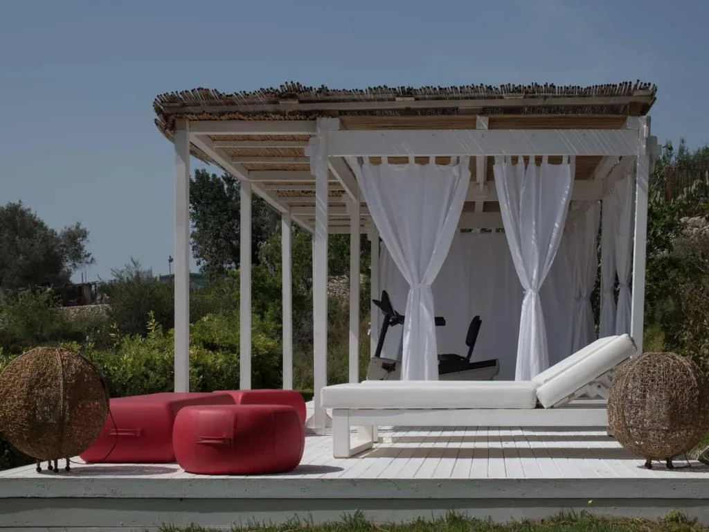 Gazebo bianco con tende e cyclette, lettino e pouf rossi su pedana di legno nel verde