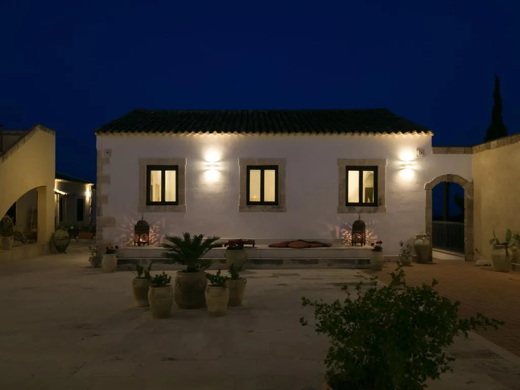 Edificio bianco illuminato di notte con cortile, fioriere e vasi in terracotta sotto cielo blu intenso
