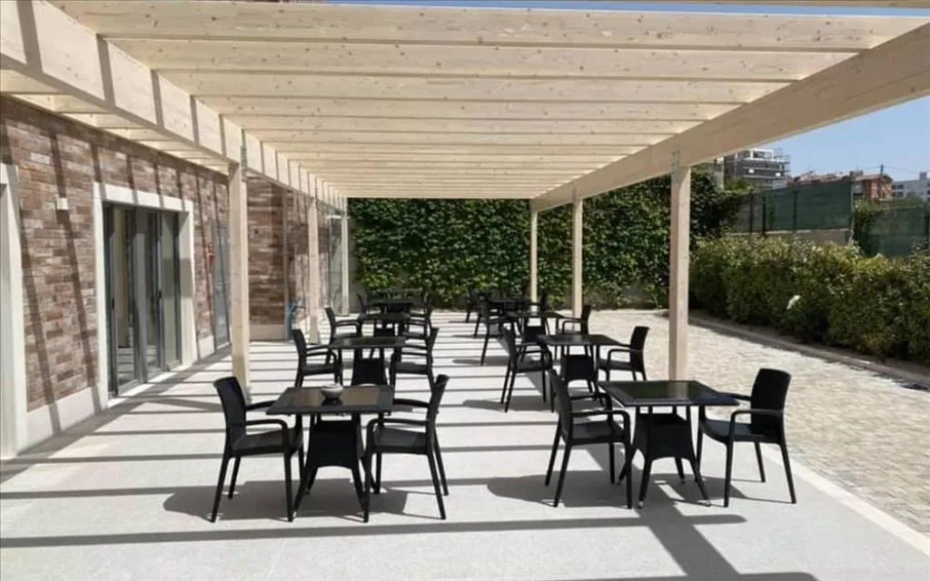 Terrazza esterna con pergolato in legno, tavoli e sedie nere lungo la siepe