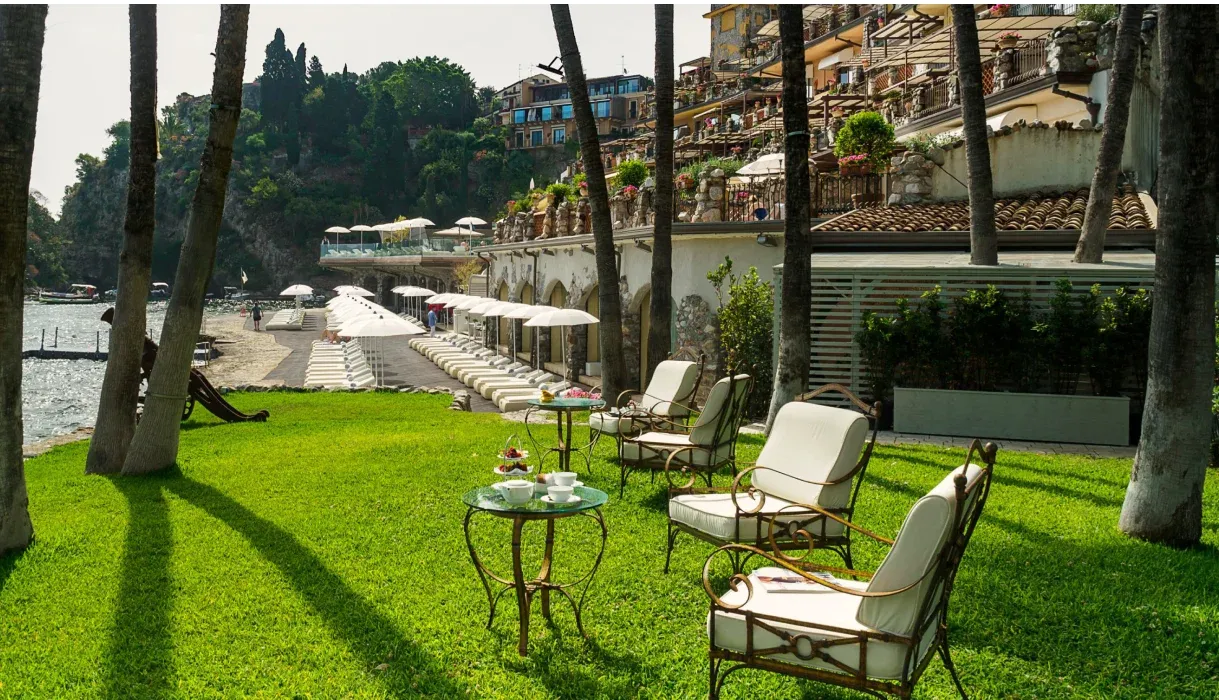 Giardino con prato, palme e poltrone affacciato sulla spiaggia con lettini e ombrelloni