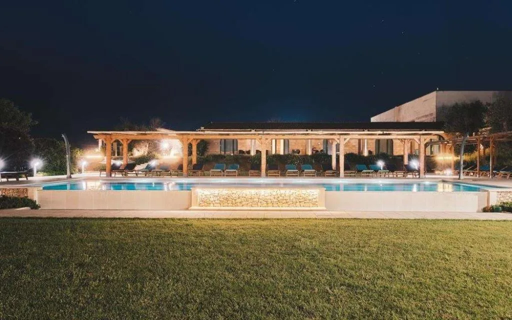 Piscina illuminata di sera con edificio in pietra e pergolati sullo sfondo