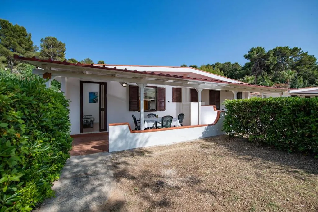 Bungalow bianco con veranda arredata con tavolo e sedie, circondato da siepi