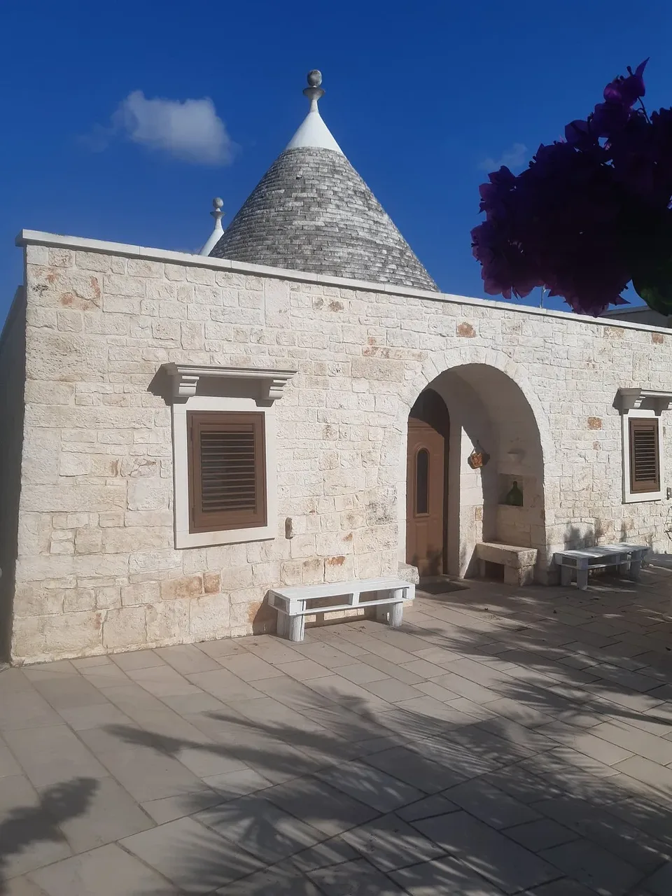 Esterno di trullo in pietra con tetto conico, cortile pavimentato e panchine bianche