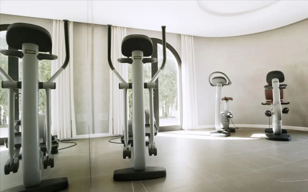 Sala fitness con attrezzi cardio e cyclette davanti a grandi finestre con tende chiare
