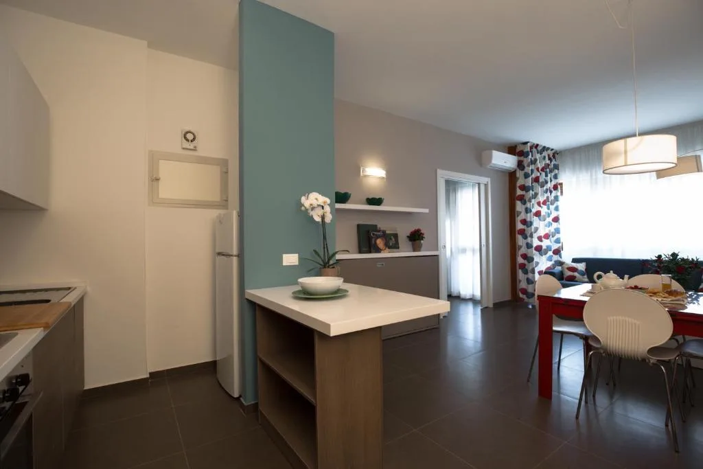 Cucina aperta con isola centrale, frigorifero e zona pranzo con tavolo rosso e sedie bianche
