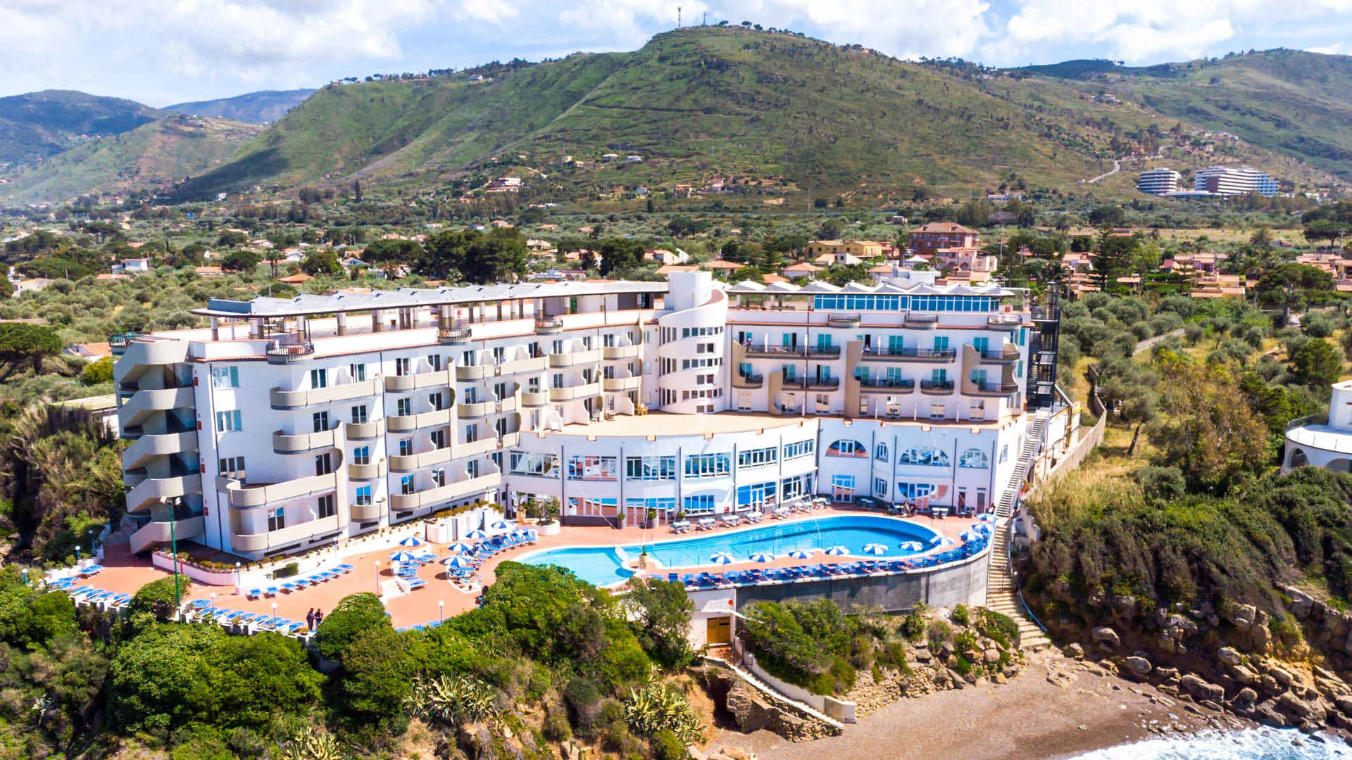 Vista aerea dell'hotel sulla costa con piscina, terrazza e colline verdi sullo sfondo