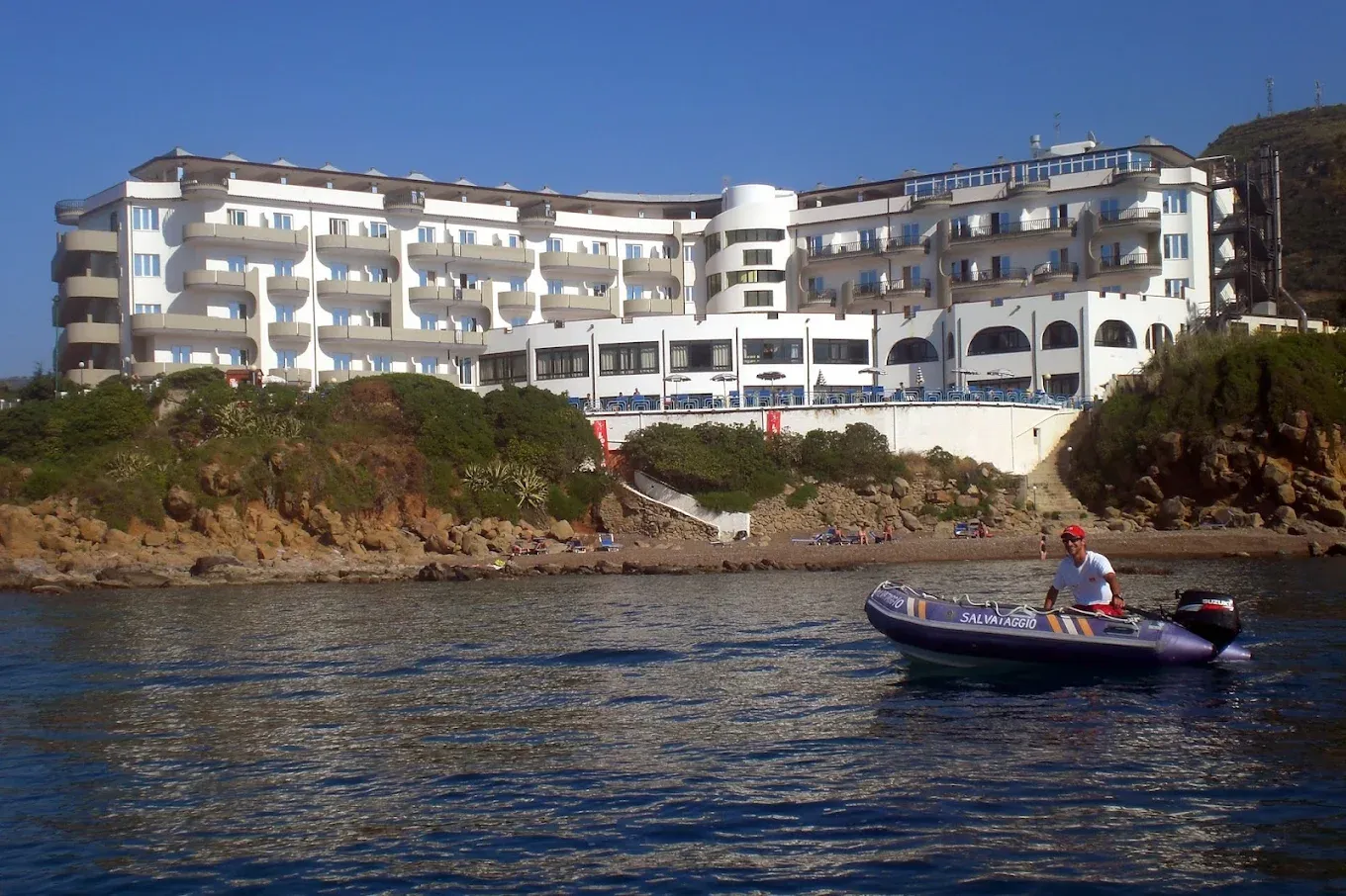 Vista dell'hotel dal mare con gommone di salvataggio e piccola spiaggia ai piedi della scogliera
