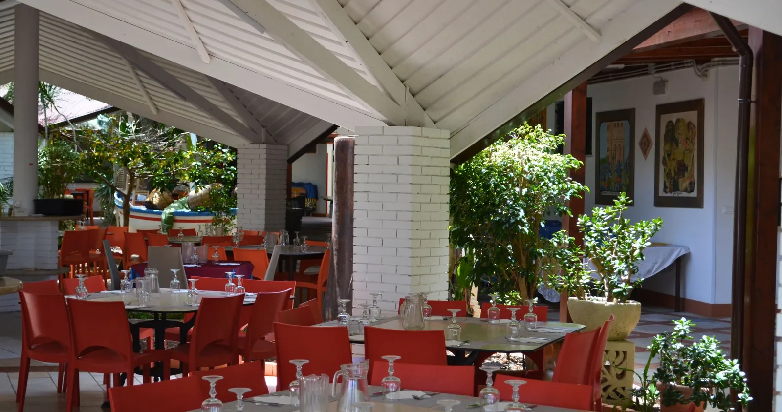 Ristorante all'aperto con sedie rosse, tavoli apparecchiati e piante verdi sotto tettoia bianca