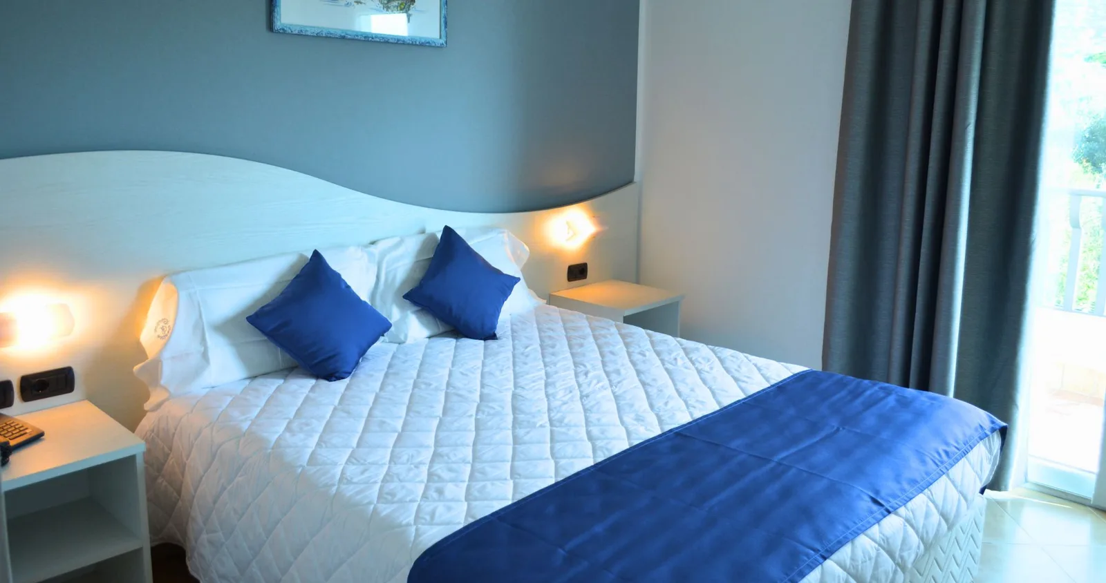 Camera matrimoniale con letto king size, cuscini blu, comodini e balcone con vista sul verde