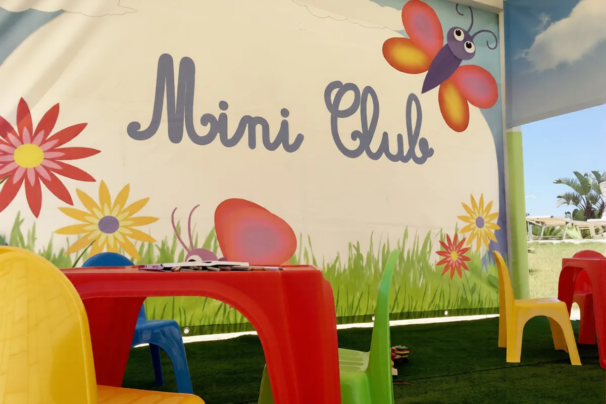 Area Mini Club per bambini con tavoli e sedie colorate e parete decorata con farfalle e fiori